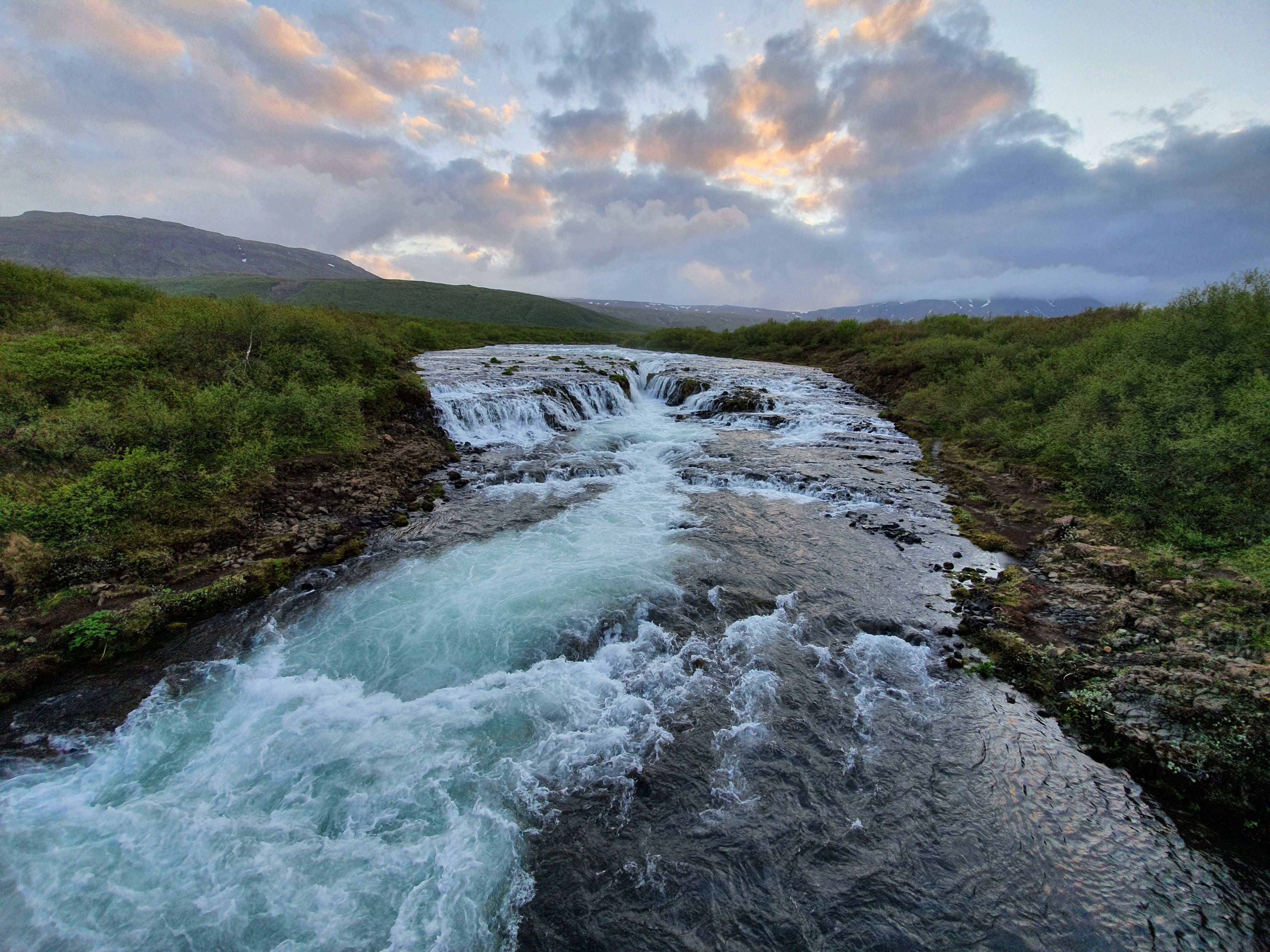 Wide view of Brúarfoss with bright turquoise water cascading over dark lava rock at sunset