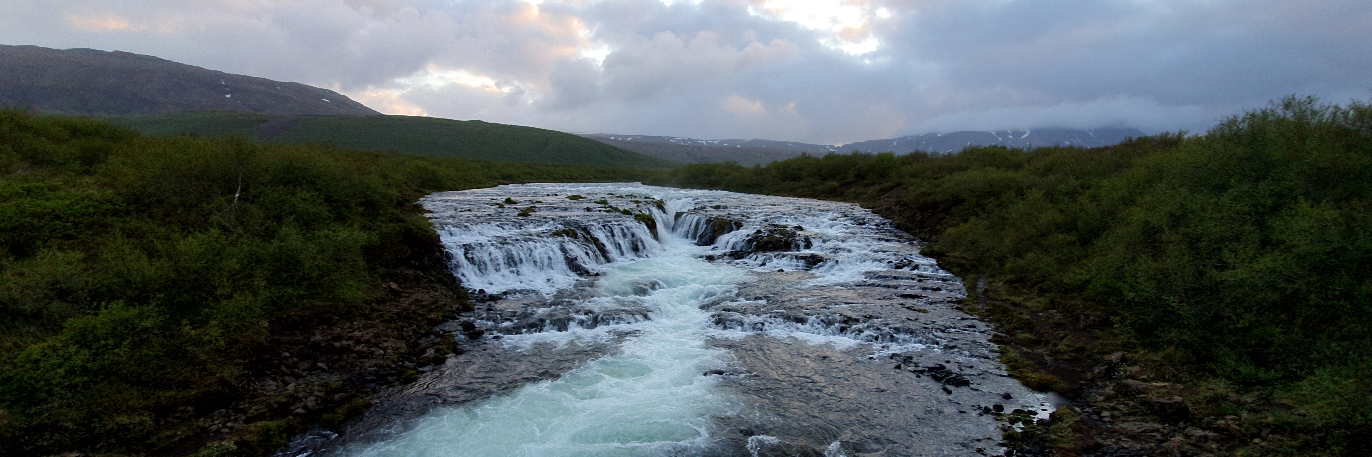 Panoramic view of Brúarfoss waterfall with green hills and cloudy sky in the background