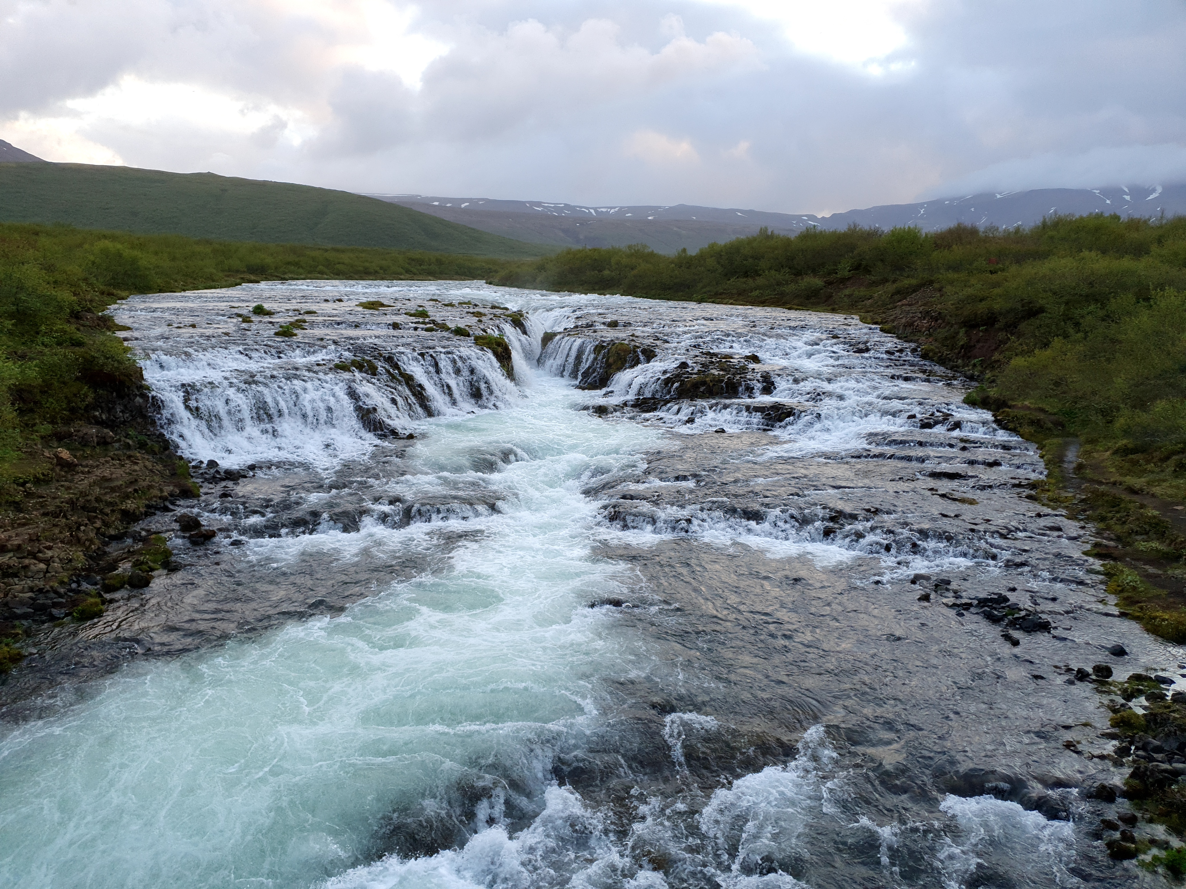 Brúarfoss waterfall close-up showing the vivid blue glacial water cascading over rocks with snow-capped mountains behind